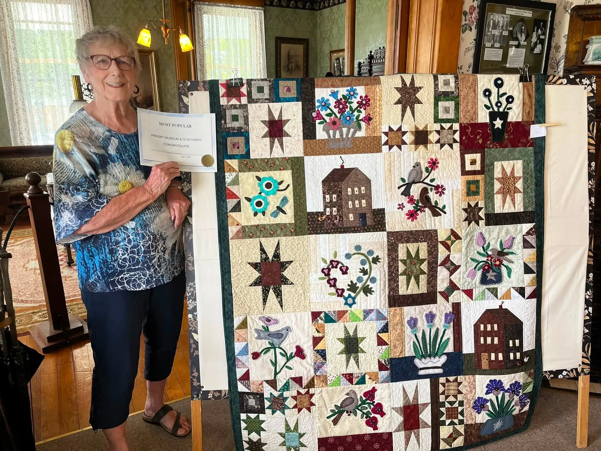 Quilter with her Honorable Mention certificate beside an appliqué sampler quilt in the museum parlor